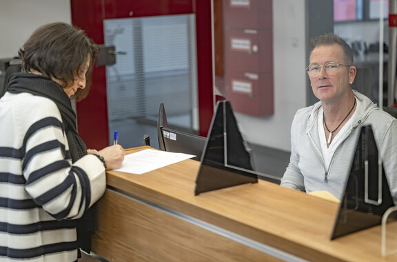Patientin steht am Empfangstresen und füllt einen Zettel aus. Hinter dem Tresen sitzt ein Mitarbeiter am Computer und schaut in die Kamera