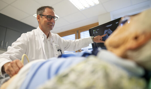 Jörg D. Fröhlich sitzt an einer Behandlungsliege und zeigt auf einen Monitor. Ein Patient liegt auf der Liege und schaut ebenfalls auf den Bildschirm und wird dabei geröntgt.