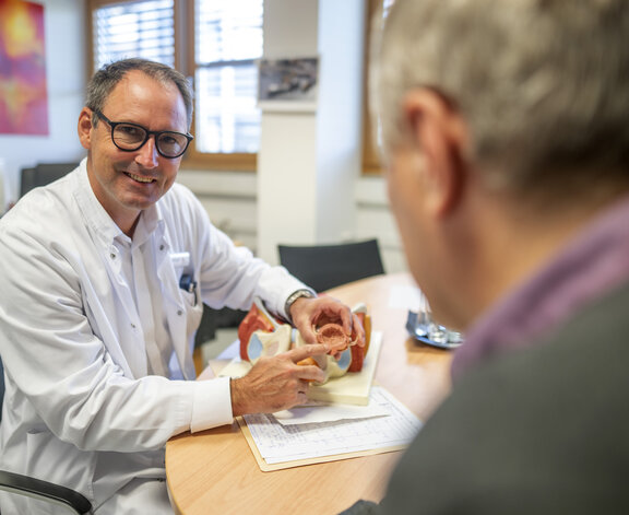 Jörg D. Fröhlich erklärt einem Patienten den urologischen Vorgang an einem Beispielkörper. Er sitzt an einem Tisch und schaut dabei dem Patienten ins Gesicht. 