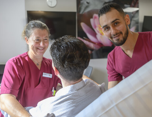 Zwei Pflegefachkräfte kümmern sich um einen Patienten, der auf einem Krankenhausbett sitzt.