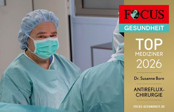 TOP Mediziner siegel 2026. Dr. med. Susanne Born steht im OP-Zimmer und schaut an der Kamera vorbei. Währenddessen operiert Sie eine Patienten. Sie trägt eine OP-Montur mit Mundschutz und Haube.