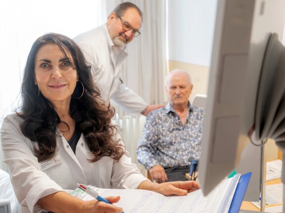 Dr. med. Frank Kuczera steht im Hintergrund neben einem sitzendem Patientem und hält diesen an der Schulter. Im Vordergrund sitzt Olga Naumov und schreibt etwas auf eine Dokument. 