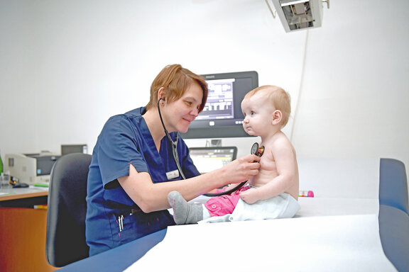 Oberärztin Susanne Schade sitzt an einer Liege und untersucht ein junges Kind, welches auf der Liege sitzt mit einem Stethoskop.