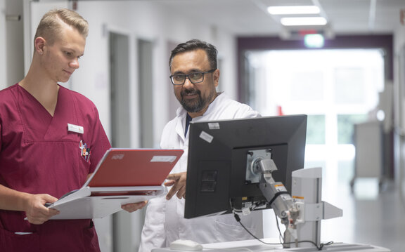 Dr. Masood steht mit einer Pflegefachkraft im Flur der Station A und schaut auf ein Tablet für die Patientenaufklärung, was die Pflegefachkraft in der Hand hält. Neben Ihnen steht ein Visitenwagen