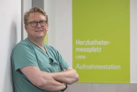 Dr. med. Frithjof Schlegel steht vor dem Eingang des Herzkatheterplatzes und der Wand und schaut in die Kamera.