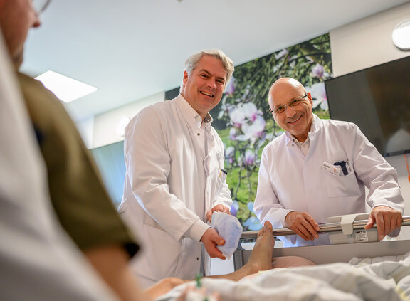 Zwei Ärzte stehen an einem Patientenbett und schauen in die Kamera. Ein Fuß der Patientin ist offengelegt
