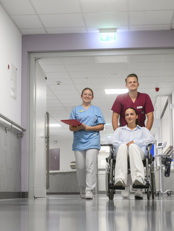 Eine Pflegekraft und eine Auszubildende als Pflegefachkraft begleiten eine Frau im Rollstuhl auf eine Station im St.-Clemens-Hospital.