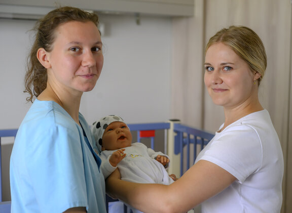 Zwei HPA Auszubildenen stehen an einem Babybett. Eine hält ein Neugeborene fest und beide schauen in die Kamera. 