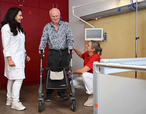 In der Klinik für Altersmedizin bespricht sich Oberärztin Olga Naumov in einem Patientenzimmer zusammen mit einer Physiotherapeutin mit einem älteren Patienten, der mit dem Gehwagen versucht zu laufen.