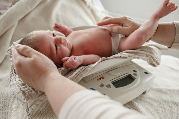 Zwei Hände halten ein Neugeborenes fest, dass auf einer Waage liegt.