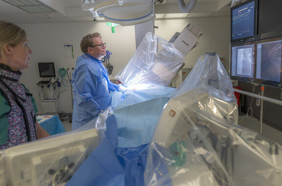 Dr. med. Frithjof Schlegel führt mit der pflegerische Leitung einen Kardiologischen Eingriff durch. Beide stehen neben der OP-Liege, welche mit OP-Decken abgedeckt ist.