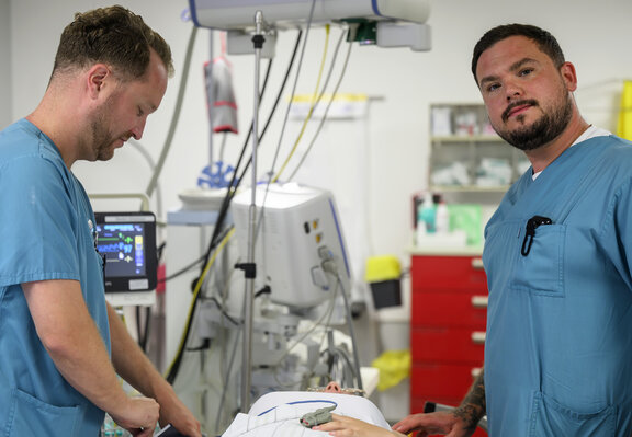 Zwei ZNA Fachkräfte stehen an einer Behandlungsliege und verkabeln einen Patienten. Einer der beiden schaut in die Kamera.