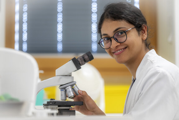 Ärztin der Gynäkologie führt eine Mikroskopuntersuchung durch. Dabei schaut sie auf und guckt in die Kamera.