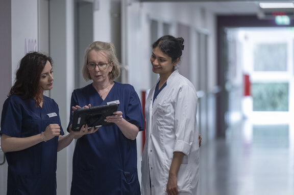 Dr. med. Ute Janßen und zwei Kolleginnen schauen auf ein IPad, dass jene in der Hand hält. Dabei stehen Sie im Flur einer Station im St.-Clemens-Hospital.