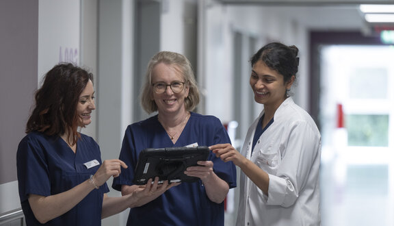 Dr. med. Ute Janßen und zwei Kolleginnen stehen im Flur. Die beiden Kolleginnen schauen auf ein IPad, das Dr. Ute Janßen in der Hand hält. Diese schaut in die Kamera.