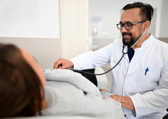 Dr. Masood untersucht Patientin mit einem Stethoskop. Dabei ist die Patientin am liegen und Dr. Masood sitzt neben der Liege auf einem Stuhl und hört die Patientin ab.
