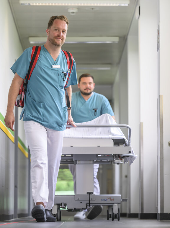 ZNA Fachkräfte schieben ein Bett durch den Flur. Einer der Fachkräfte hat ein Rettungsrucksack auf. Beide schauen in die Kamera.
