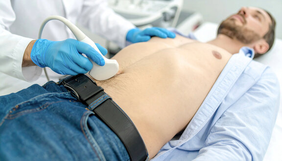 Ein Mann liegt mit aufgeknöpftem Hemd und freiem Oberkörper auf einer Untersuchungsliege. Ein Arzt macht eine Ultraschalluntersuchung von seinem Bauch.
