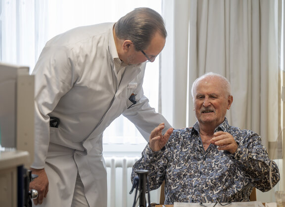 Dr. med. Frank Kuczera steht mit einem weißen Kittel vor einem Patienten, der im Stuhl sitzt, und legt seine Hand kümmernd auf seine Schulter. Der Patient hat seine Hände leicht angehoben. 