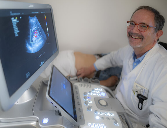 Dr. med. Andreas Fußhöller sitzt am Computer und schaut sich Röntgenergebnisse an. Im Hintergrund sieht man einen Patienten, der auf einer Liege liegt und geröntgt wird. Dr. med. Andreas Fußhöller lächelt in die Kamera.