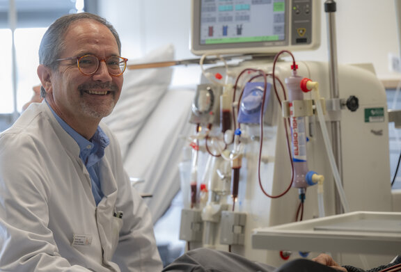Dr. med. Andreas Fußhöller sitzt vor einem Patienten der im Krankenbett liegt und lächelt freundlich in die Kamera. 