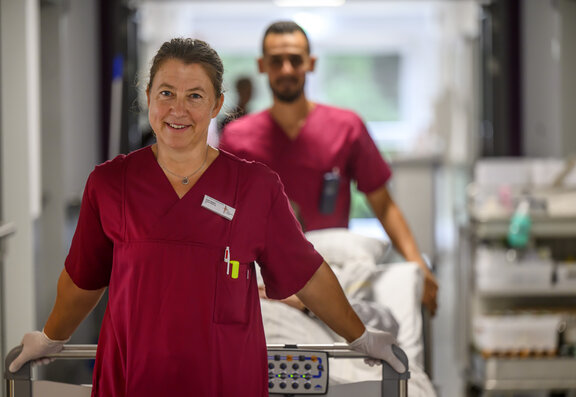 Zwei Pflegefachkräfte schieben ein Bett durch den Flur. Auf der rechten Seite steht ein Versorgungswagen.