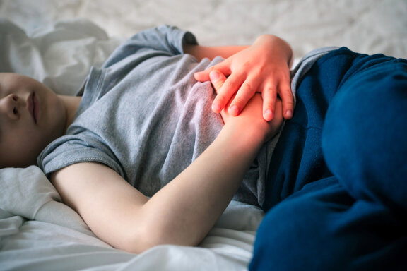 Bild eines Kindes, dass mit Bauchschmerzen im Bett liegt. Der Bereich am Bauch, der das Kind umfasst ist mit rot hervorgehoben.