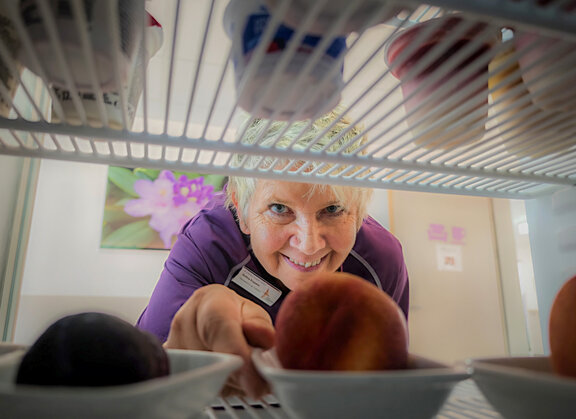 Mitarbeiterin der Cafeteria schaut in einen Kühlschrank und holt eine Schale raus. Das Bild ist aus dem Kühlschrank heraus gemacht und die Mitarbeiterin schaut in die Kamera. 