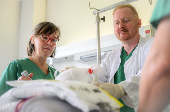 Dr. Tom Edwin Mccracken und eine HPA führen gemeinsam eine Simulation durch. Die HPA schaut dabei zu wie Dr. Tom Edwin Mccracken ein Beatmungsgerät stabilisiert und hält dabei ein Schlauch in der Hand.