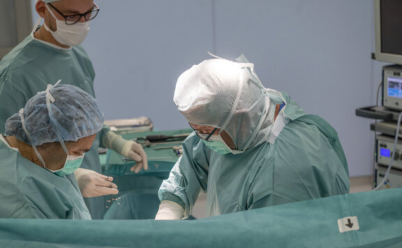 Dr. med. Susanne Born und Dr. Wolf sind in einem OP-Raum und operieren einen Patienten. Ein weiterer OTA steht im Hintergrund und bringt ein Operationswerkzeug.