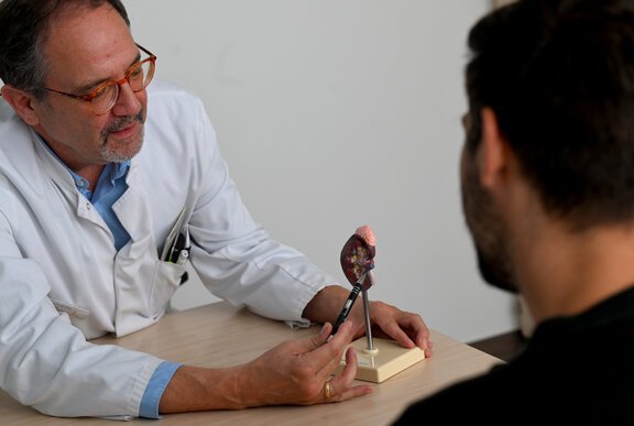 Dr. med. Andreas Fußhöller zeigt einem Patienten, von dem man nur die Haare sieht, ein Modellherz mit seinem Kugelschreiber. Das Modellherz ist mit einer Metallstange auf einem Holzblock befestigt.