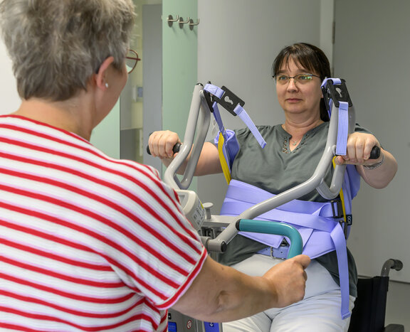 Pflegeüberleitung steht mit Patientin, die gerade eine Übung macht, an einem Geriatrischem Gerät. 