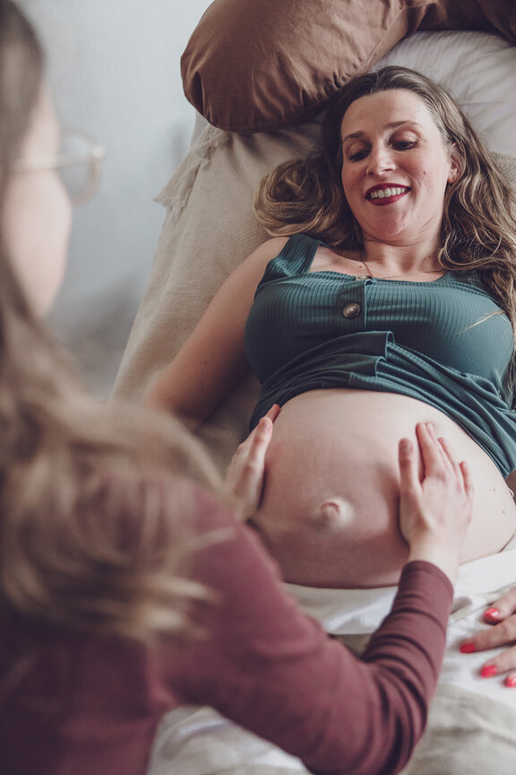 Hebamme untersucht eine schwangere Frau, indem Sie den Bauch abtastet. Die Patientin liegt auf einer Liege und schaut zu, wie die Hebamme den Bauch abtastet. 