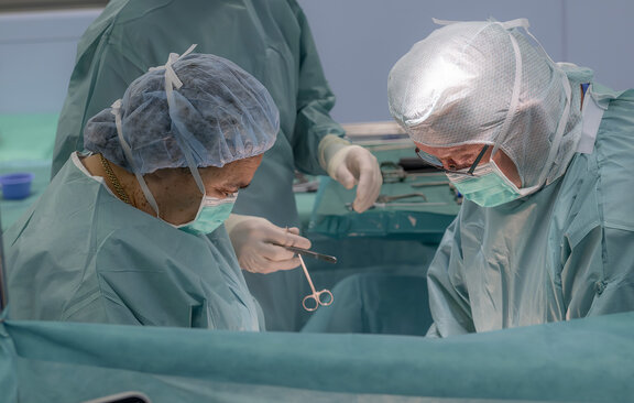 Dr. med. Susanne Born und Dr. Wolf sind in einem OP-Raum und operieren einen Patienten. Ein weiterer OTA steht im Hintergrund und bringt ein Operationswerkzeug.