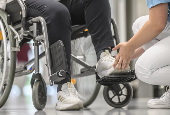 eine Pflegekraft setzt den Fuß eines Patienten auf die Fußstütze eines Rollstuhls