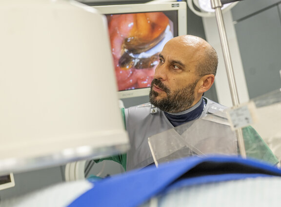 Dr. med. Kerem Bulut steht im endoskopischen Behandlungsraum und betrachtet Bildschirme. Auf dem hinter ihm ist ein Teil des Darms zu sehen.