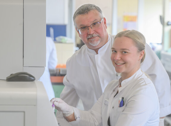 Laborleitung Massimo Battistuzzi und eine Labor-Fachkraft stehen vor einem Lasergerät und schauen in die Kamera. Beide tragen einen weißen Kittel und die Fachkraft hält eine Probe in der Hand.