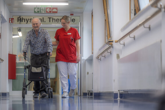 Ergotherapeutin läuft mit älteren Patientin, die einen Rollator hat, durch den Flur der Geriatrie.
